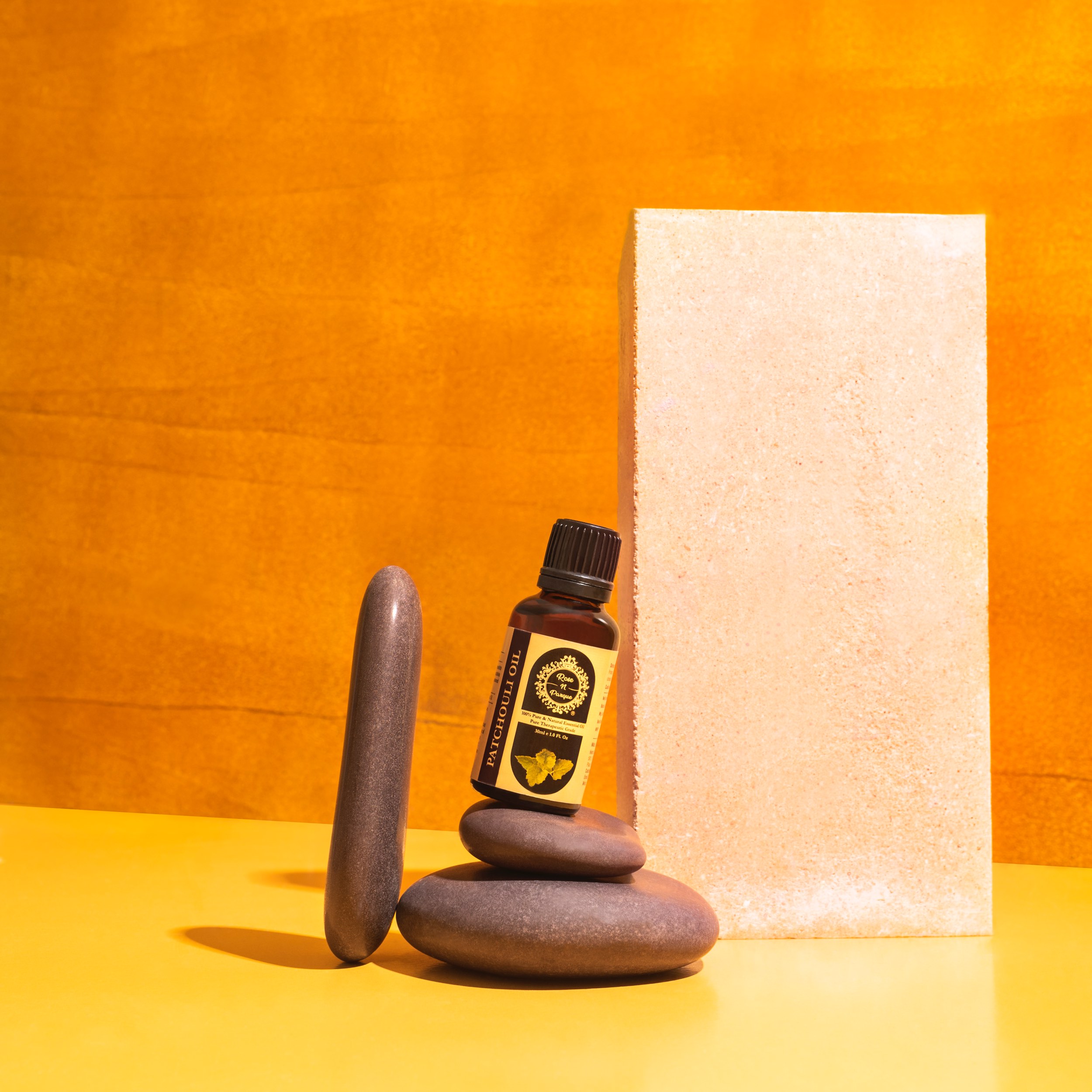 Brown essential oil bottle with a black cap placed among smooth stones and a textured block against a yellow background.