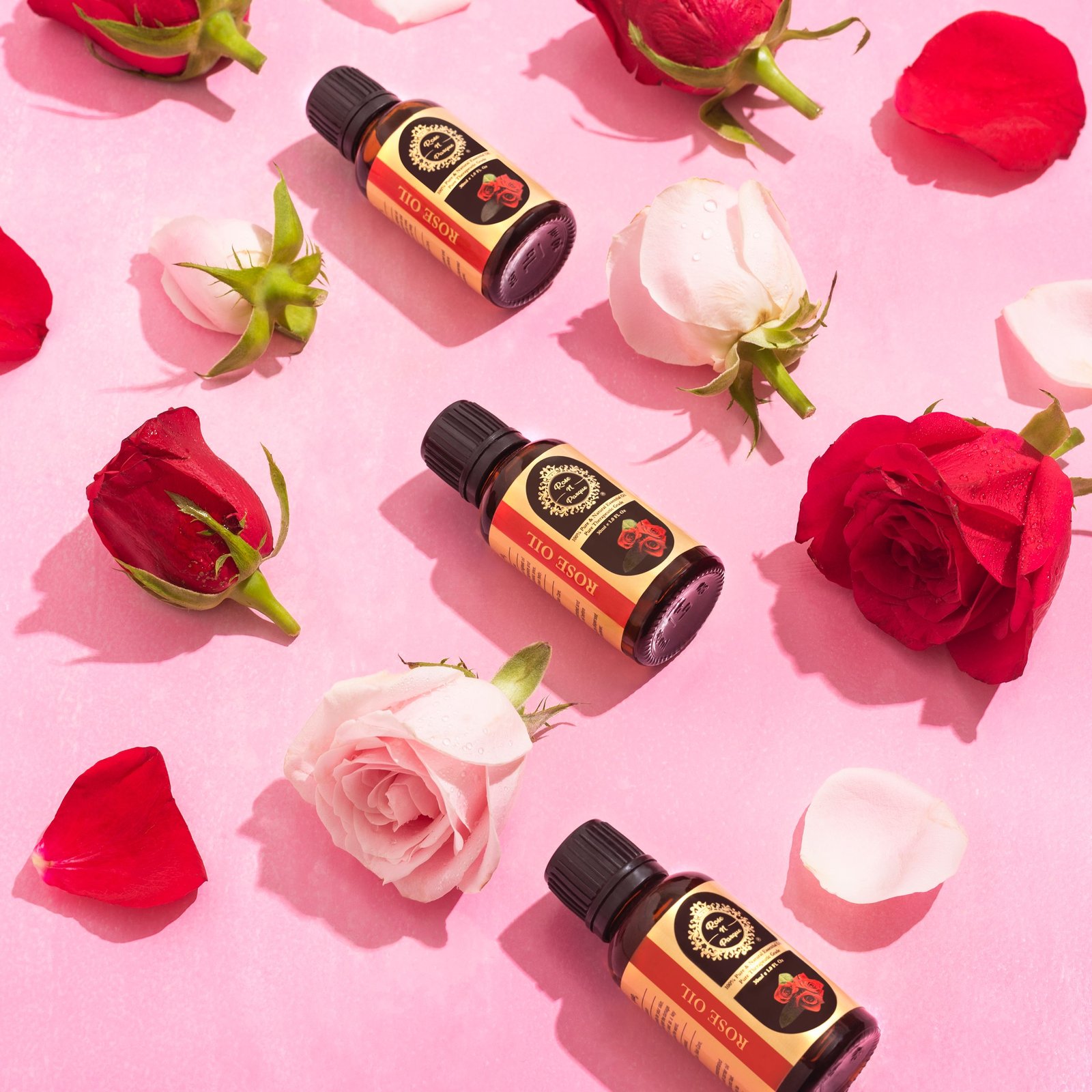 Four bottles of rose oil surrounded by pink and red rose petals on a pink background.