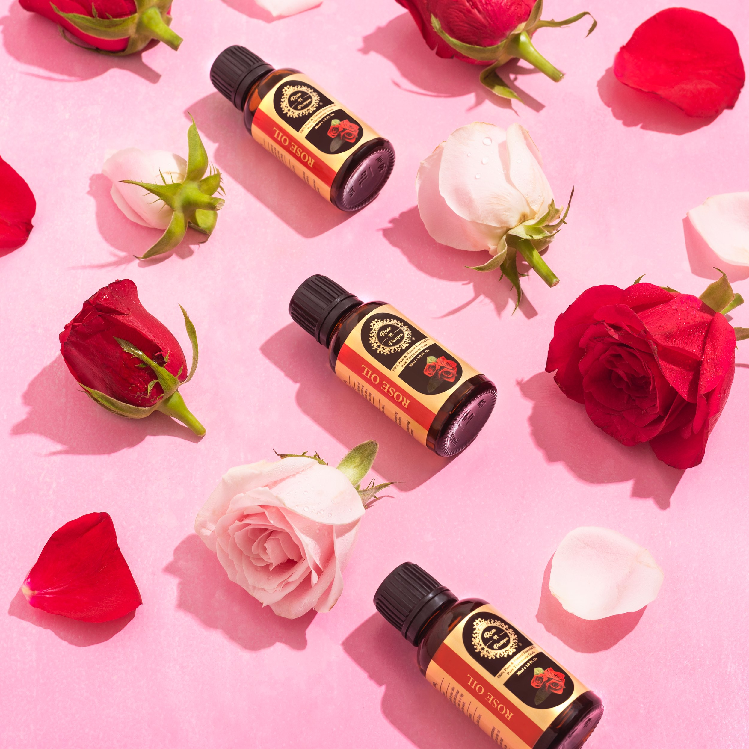 Four bottles of rose oil surrounded by pink and red rose petals on a pink background.