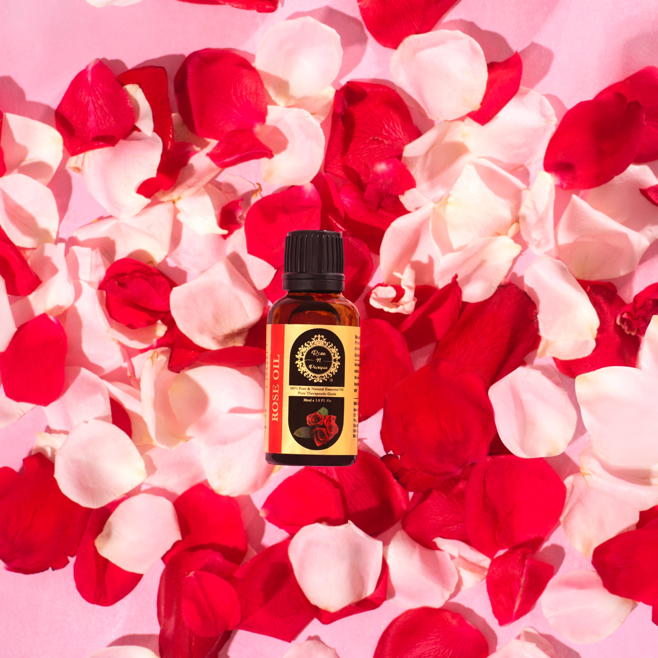 Brown bottle of rose oil surrounded by red and white rose petals on a pink background.