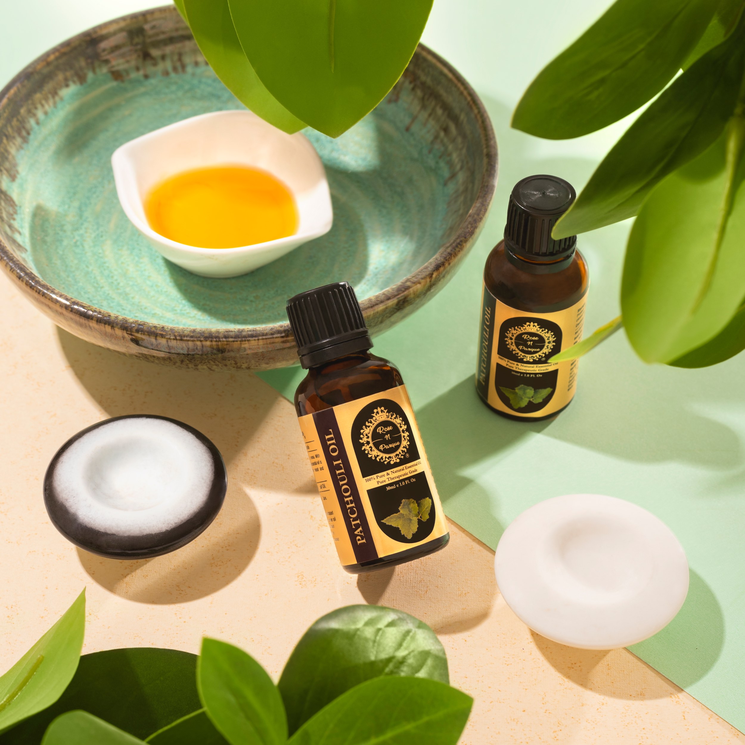 Two bottles of patchouli oil placed near a small bowl of oil and surrounded by green leaves.