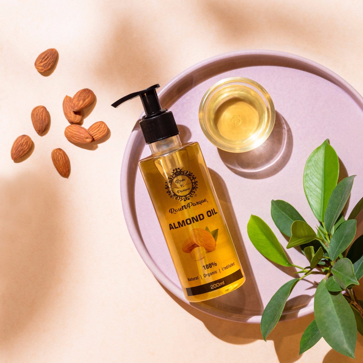 Bottle of 100% almond oil with a pump dispenser next to a small glass and almonds on a light surface.