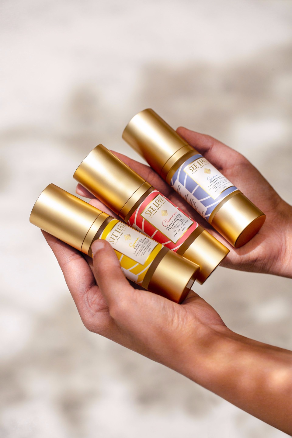 Three skincare products held in hands, featuring gold packaging and colorful labels.