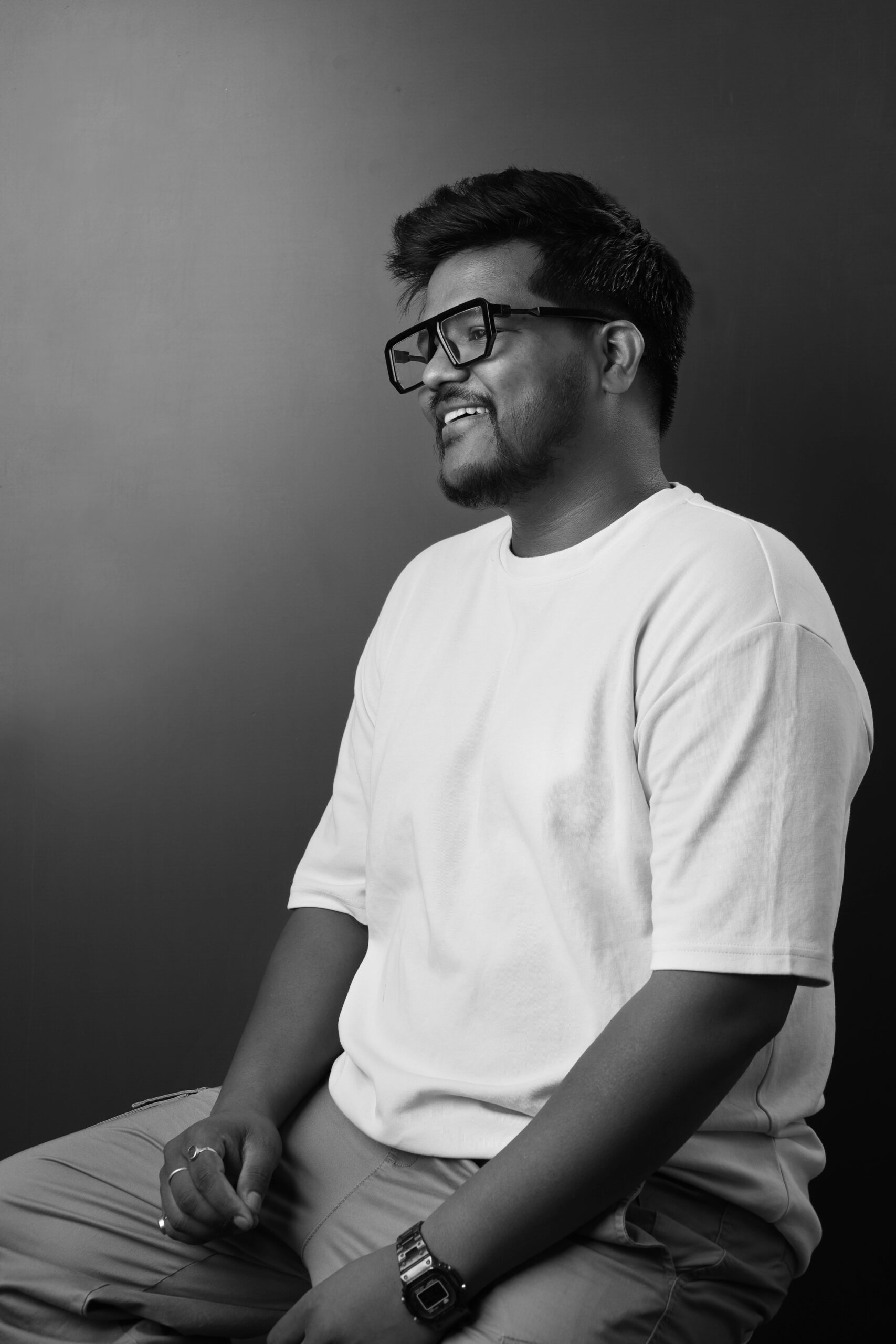 A man wearing glasses and a white t-shirt, smiling while seated against a dark background.