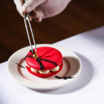A red macaron being placed on a plate with tongs, featuring cream filling and decorative elements.