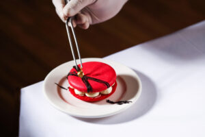 A red macaron being placed on a plate with tongs, featuring cream filling and decorative elements.