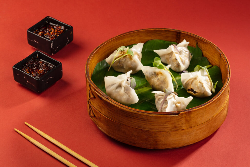 A bamboo steamer filled with steamed dumplings, accompanied by dipping sauces in black bowls and chopsticks on the side.