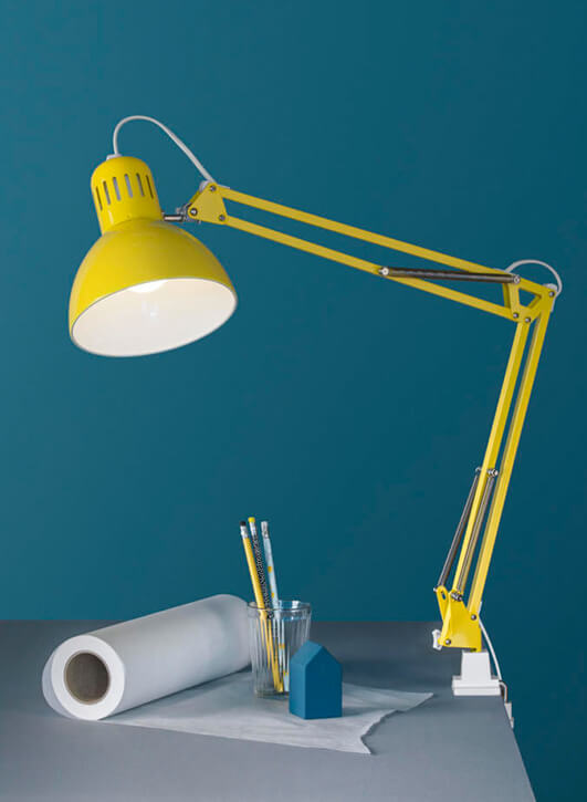 Yellow desk lamp with an adjustable arm, glass cup with pencils, and a blue house-shaped object.