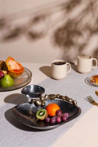 A black plate with assorted fruits including kiwi, grapes, and an orange, accompanied by cups and a decorative bowl on a table.