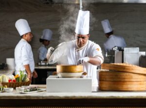 Chef in a white uniform lifting a lid off a bamboo steamer in a kitchen with steam rising.