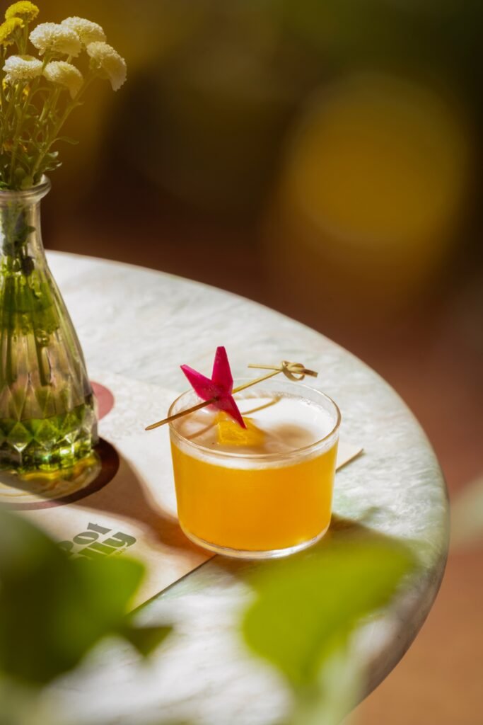 A yellow citrus cocktail with a red star garnish sits beside small white flowers on a sunlit marble table.