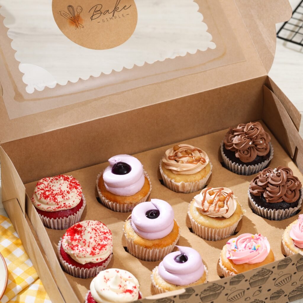 An assortment of frosted cupcakes fills a kraft bakery box, with red velvet, chocolate, and pastel toppings.