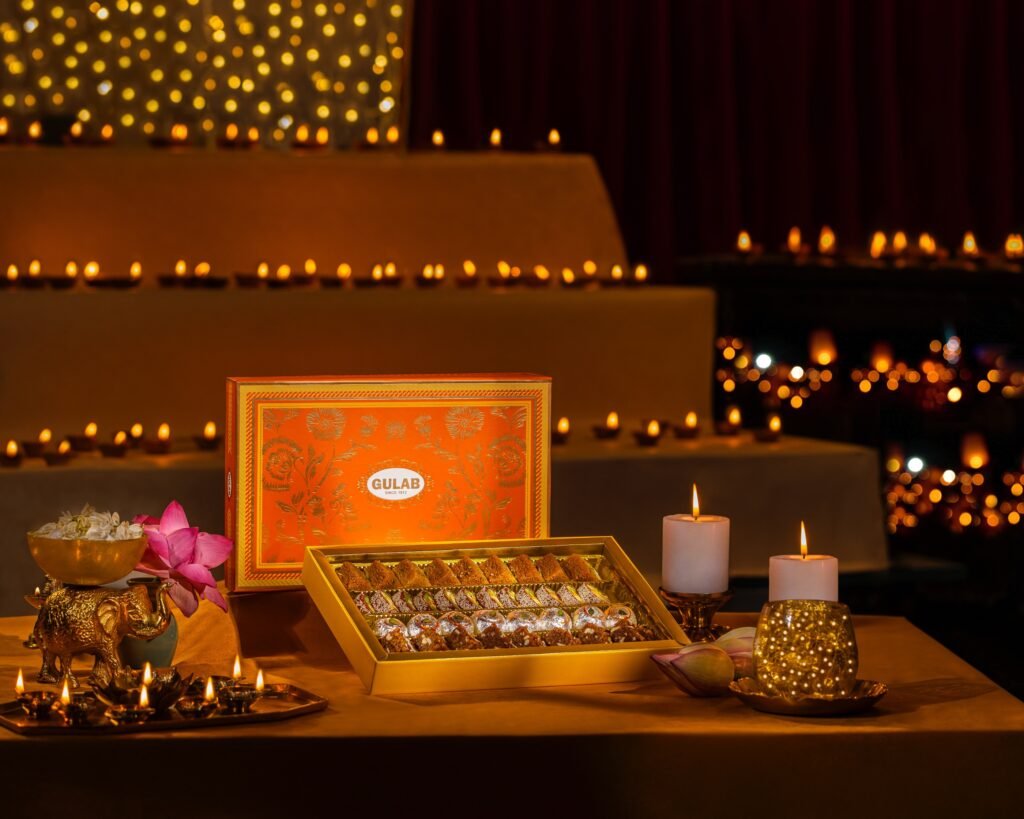 Rows of assorted Indian sweets sit in an open Gulab gift box beside lotus flowers and glowing diyas at a festive display.