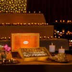 Rows of assorted Indian sweets sit in an open Gulab gift box beside lotus flowers and glowing diyas at a festive display.