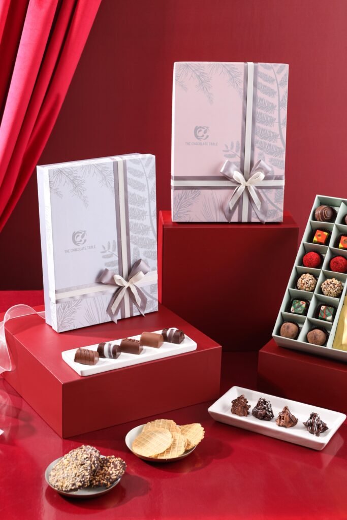 Ribbon-tied The Chocolate Table gift boxes stand among plated chocolates, truffles, wafers, and cookies on red display blocks.