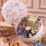 A floral keepsake hamper displays chocolates, sweets, flowers and a candle, with bowls of dates, biscuits and blue candies nearby.