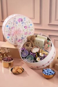 A floral keepsake hamper displays chocolates, sweets, flowers and a candle, with bowls of dates, biscuits and blue candies nearby.
