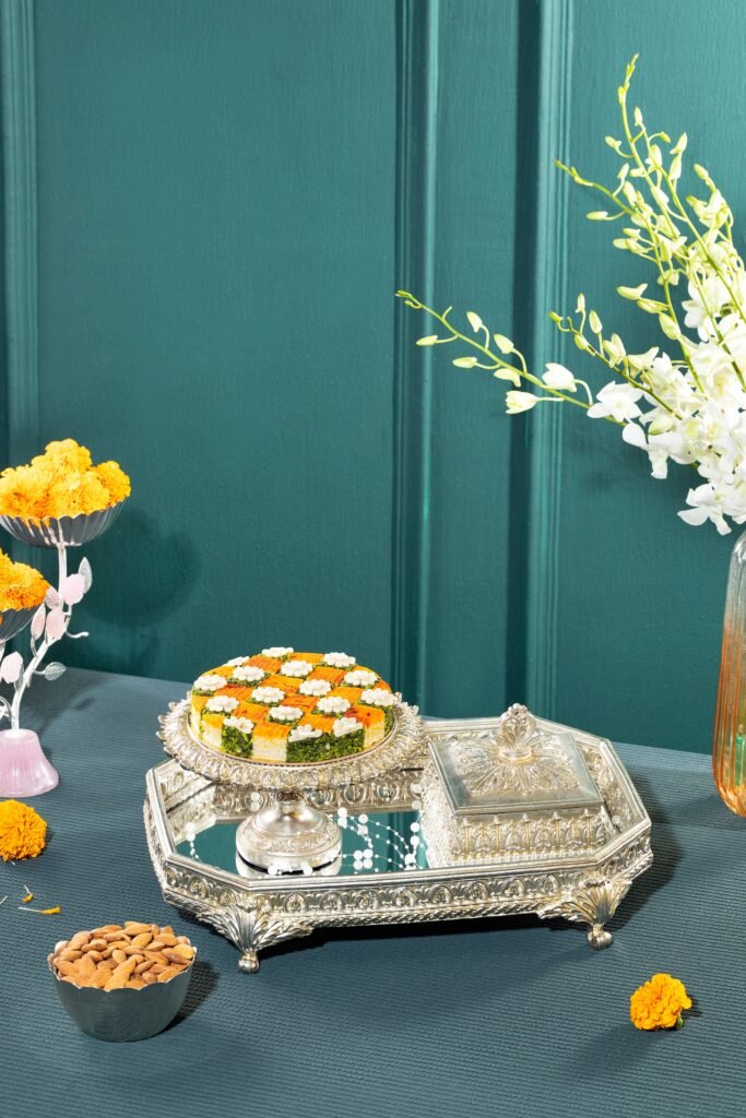 A layered savory cake topped with piped cream sits on an ornate silver tray beside almonds, marigolds, and white flowers.