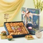 An ornate blue sweets gift box filled with assorted Indian mithai, cookies, and savory snacks beside flowers.