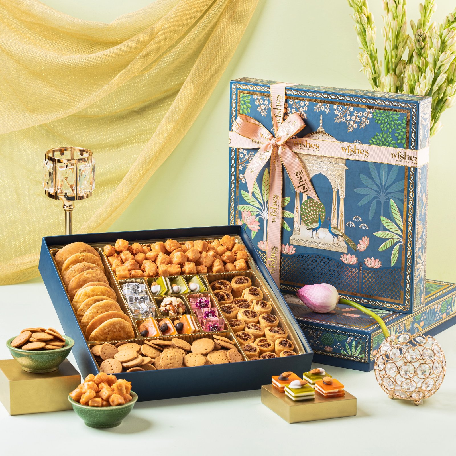 An ornate blue sweets gift box filled with assorted Indian mithai, cookies, and savory snacks beside flowers.