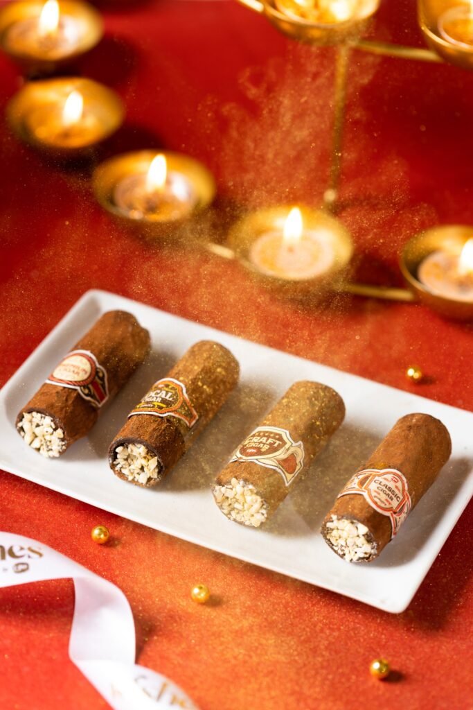 Four branded cigars rest on a white platter beside glowing tealight candles and gold accents on red fabric.